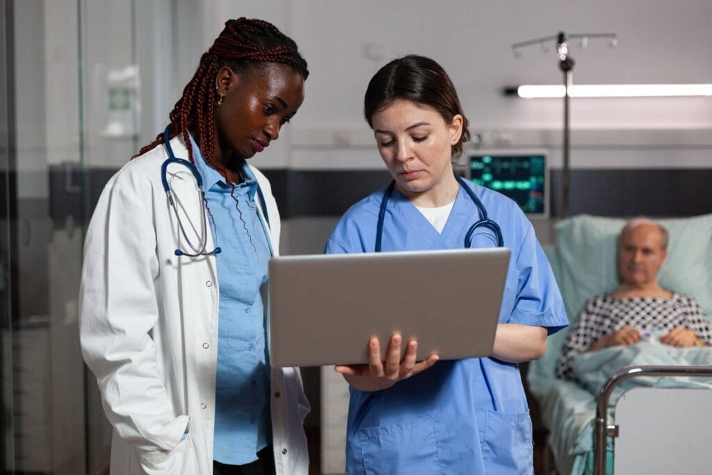 Two nurses utilizing a laptop in a hospital room while discussing the importance of a local health insurance agent.