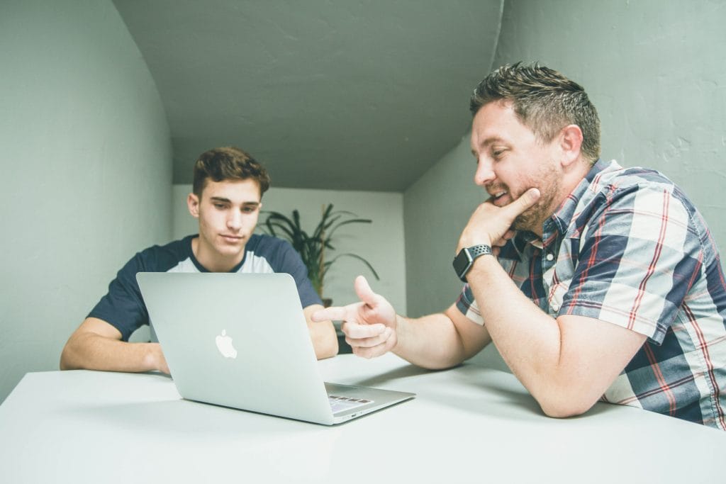 Health insurance broker consulting with a small business owner at a table. The broker, a middle-aged man in a suit, is explaining insurance options, while the business owner, a younger man in business casual attire, listens attentively. Documents and a laptop are on the table, emphasizing a professional consultation.