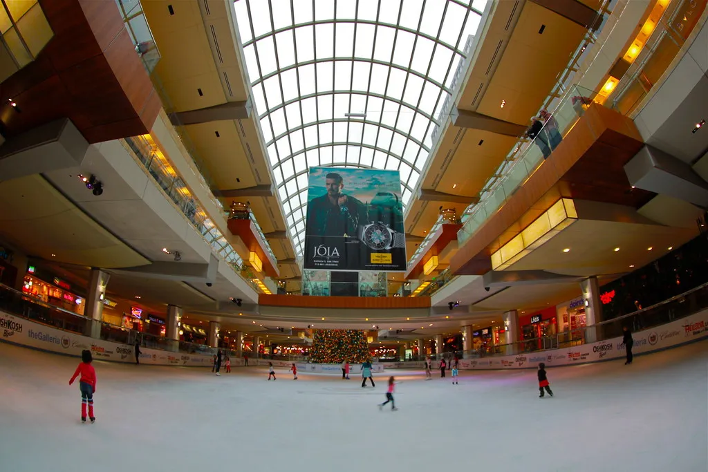 Interior view of a shopping mall with a large ice skating rink, featuring people skating under a high arched glass ceiling, surrounded by multiple stores and a large advertisement banner. Suitable for articles on shopping centers, recreational activiti